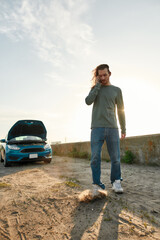 Full length shot of young man talking on the phone with car service, assistance or tow truck while...