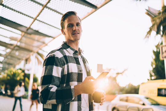 Portrait Of Handsome Caucasian Millennial Male Listening Music During Free Time On Sunny Day In City, Young 20s Hipster Guy Holding Smartphone Enjoying Music Via Earphones Connected To Smartphone