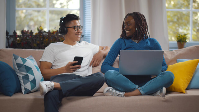 Happy young diverse couple relaxed at home on couch surfing internet on mobile phone and using laptop