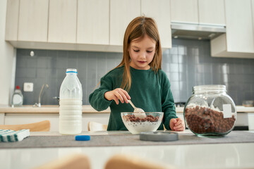 A cute girl prepearing her quick breakfast made of chocolate cereal balls and milk stirring with a...