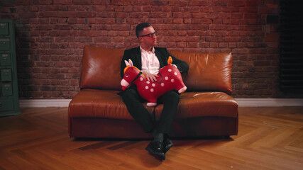 Obraz premium Portrait of young man in eyeglasses and suit sitting on leather coach spanking inflatable red horse