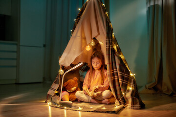 A cute girl wearing pyjamas sitting on a floor cross-legged barefoot in a self-made hut made of a plaid holding and scrolling a book with fancy garlands all around the hut