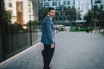 Naklejka premium Successful businessman dressed in trendy jacket looking at camera while walking in downtown, full length portrait of mature male entrepreneur 40 years old in formal wear posing at urban street