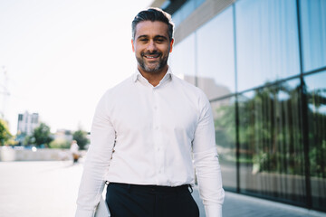 Half length portrait of cheerful businessman 40 years old dressed in white shirt smiling at camera in financial district, happy Caucasian male employer in formal clothes posing in city downtown
