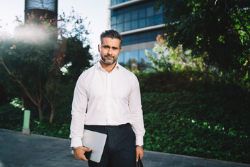 Fototapeta premium Half length portrait of successful businessman dressed in formal white shirt looking at camera at sunny city street, middle aged Caucasian employer with modern netbook technology posing outdoors