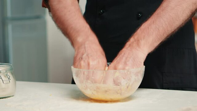 Senior chef sprinkling flour and kneading dough in glass bowl. Close uRetired elderly baker with bonete and apron, spreading, sieving sifting raw ingredients by hand baking homemade pizza, bread.