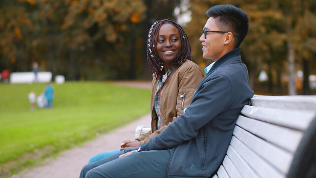 Flirting diverse couple sitting on bench in park spending time together - Powered by Adobe