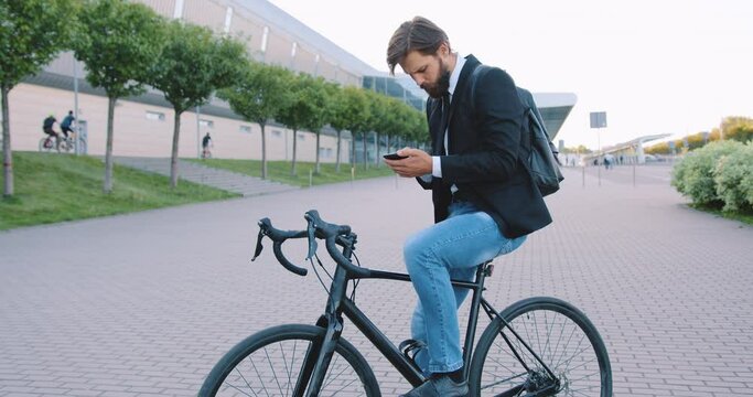 Attractive stylish young bearded man sitting on bike during stop on the pedestrian path on the city building background in daytime,front view