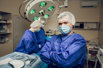 Portrait of a surgeon in a protective mask in the operating room. Behind him, two other doctors are preparing for surgery