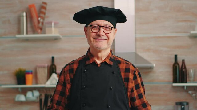 Caucasian old man wearing apron in home kitchen smiling at camera. Retired elderly baker with bonete preparing pastry ingredients on wooden table ready to cook homemade tasty bread, cakes and pasta.