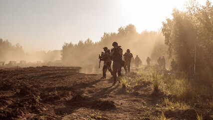 A group of soldiers goes through the field along the forest in the smoke. - obrazy, fototapety, plakaty