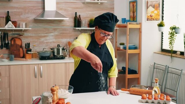 Elderly woman adding flour on table by hand sitting in the home kitchen. Retired senior baker with bonete and uniform sprinkling, sifting, spreading rew ingredients baking homemade pizza and bread.