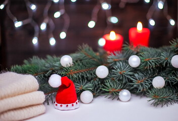 Christmas composition with Santa's hat, fir branches, white decorations on a blurry background with red candles and garland lights.