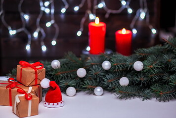 Christmas composition with Santa's hat and gifts, fir branches, white decorations on a blurry background with red candles and garland lights.