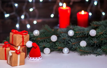 Christmas composition with Santa's hat and gifts, fir branches, white decorations on a blurry background with red candles and garland lights.