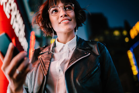 Prosperous party girl with smartphone technology enjoying nightlife in metropolitan city, happy millennial in optical eyewear and modern headphones listening funny music set during evening walk