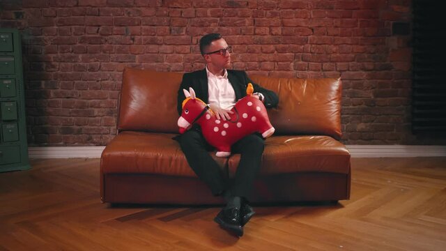 Portrait of young man in eyeglasses and suit sitting on leather coach spanking inflatable red horse