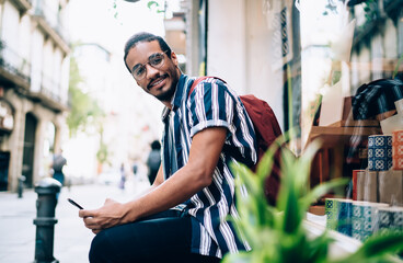 Half length portrait of smiling dark skinned male millennial using smartphone communicating and browsing on free time, smiling african american looking at camera satisfied with 4G connection on street