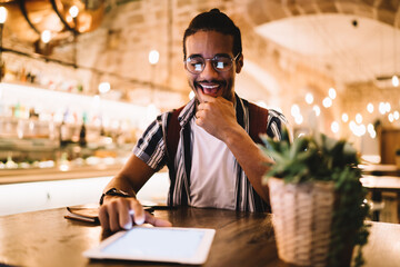 Happy dark skinned male 20s sitting in cafe interior watching video on modern digital tablet, smiling african american hipster guy excited with new game on touchpad using 4G internet connection