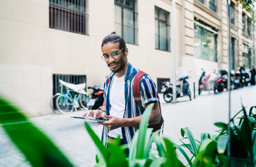 Positive dark skinned male in casual wear and spectacles walking on city street holding modern touchpad,smiling handsome hipster guy searching information on navigating via app on digital tablet