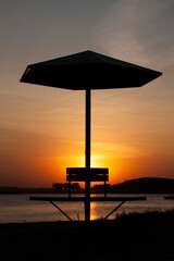 landscape photography of a silhouette of an isolated stationary beach umbrella with round bench on the lake shore in the first plan and calm lake during beautiful orange autumn sunset