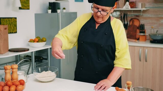 Woman chef taking wheat flour from glass bowl and sieving on table. Retired senior baker with bonete and uniform sprinkling, sifting, spreading rew ingredients baking homemade pizza and bread.