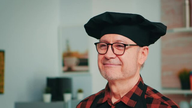 Elderly age man wearing chef bonete smiling in home kitchen. Close up portrait of happy retired old baker with glasses and apron looking at camera ready to cook homemade pastry with flour and eggs.