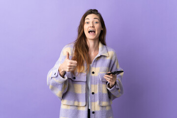 Young Ireland woman isolated on purple background using mobile phone while doing thumbs up