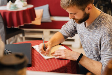 A bearded man sits at a table in a cafe and writes in a notebook.