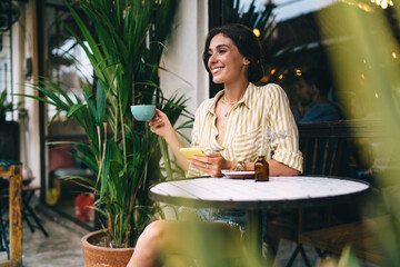 Happy young ethnic woman with cup of coffee sitting on terrace