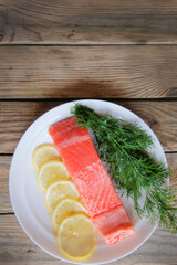 A piece of fresh trout fillet with lemon and dill greens on a white plate on a wooden table waiting for cooking. Restaurant menu. Dietary product. Keto diet. The concept of healthy eating.