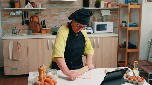 Senior woman with bonete looking recipe at the digital laptop. Retired lady following culinary advice on tablet, learning cooking tutorial on social media, using wooden rolling pin forming the dough.