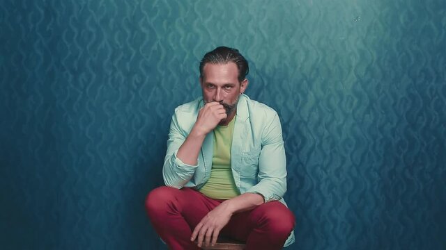 Old beared man sitting on the wooden chair on the blue background. Man show somebody middle finger. Mad aggressive man. High quality FullHD footage