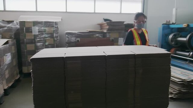 Asian woman factory worker doing arrangement cardboard paper to be stack for preparation process in warehouse room. Asia female wearing face mask while working in factory. New normal wear of people.