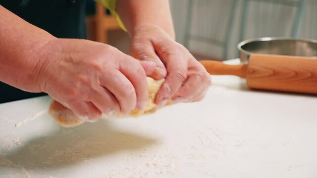 Close up of grandmother hands kneading on table in home kitchen. Retired elderly baker with bonete mixing ingredients with sifted wheat flour kneading for baking traditional cake and bread.