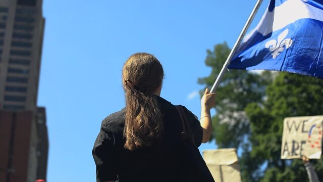 Rear view of young woman standing, holding quebec country flag and waving it in the air during protest against fake news with crowd holding banner