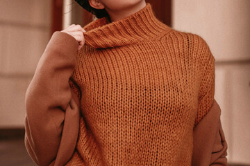 Young woman with cropped face in the orange knitted sweater and brown coat, outdoor portrait in daylight. Cozy autumn.