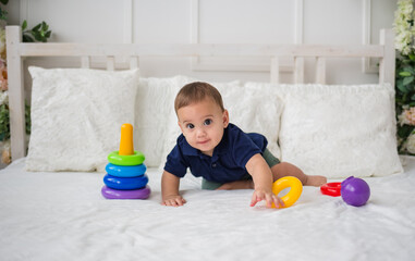 funny baby boy looks at the camera and plays with a pyramid toy on a white bed