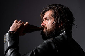 Brutal bearded biker in leather jacket with bottle of beer. Handsome man with beer. Pensive, cool, shadows, Bottle of beer.