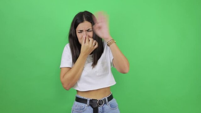 Repulsion to bad smell. Young beautiful woman over isolated green background clutching her nose with her fingers, holding her breath to avoid the stench, the intolerable smell, showing a stop gesture.