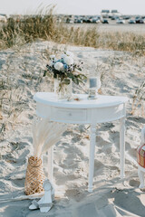 Maritime wedding bride getting ready decoration on the northsea beach, vintage boho white table, bouquet in vase, big soft blue candle, rings on shell, shoes on the sand.