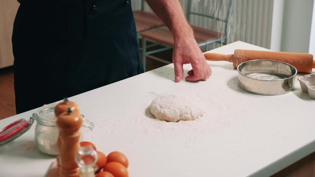 Close up of retired baker pouring wheat flour on dough for bread. Elderly senior baker with bonete and uniform sprinkling, sifting, spreading rew ingredients by hand, baking homemade pizza and cookies