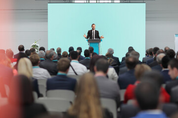 People sitting in a conference hall and a man in a suit giving a speech