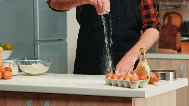 Mature old baker wearing apron using flour for food recipe. Retired senior chef with bonete and uniform sprinkling, sieving sifting raw ingredients by hand baking homemade pizza and bread.