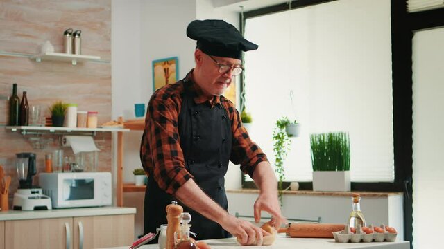 Confectioner working at home with raw dough in modern kitchen recording the recipe. Retired elderly baker with bonete mixing ingredients with sifted flour kneading for baking traditional bread.