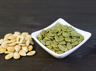 Bowl of tasty and healthy roasted pumpkin seeds with heap of raw seeds isolated on wooden table
