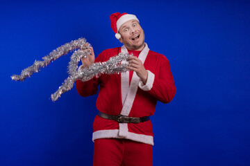 Male actor in a suit and hat of Santa Claus with a silver tinsel garland dancing and posing on a...