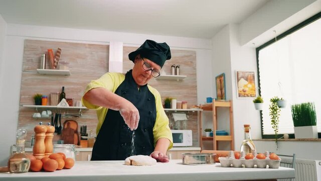 Woman elderly cook sprinkling a piece of dough with flour. Retired senior baker with bonete and uniform sprinkling, sifting, spreading rew ingredients on dough, baking homemade pizza and bread.