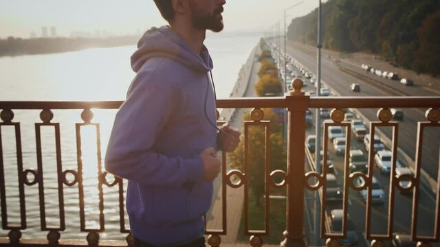 Urban training. Young man running on bridge above highway early in morning, side view, slow motion