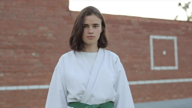 Portrait of a young karate girl in a white kimono who stands against a brick wall background. Slow motion. Blurred background